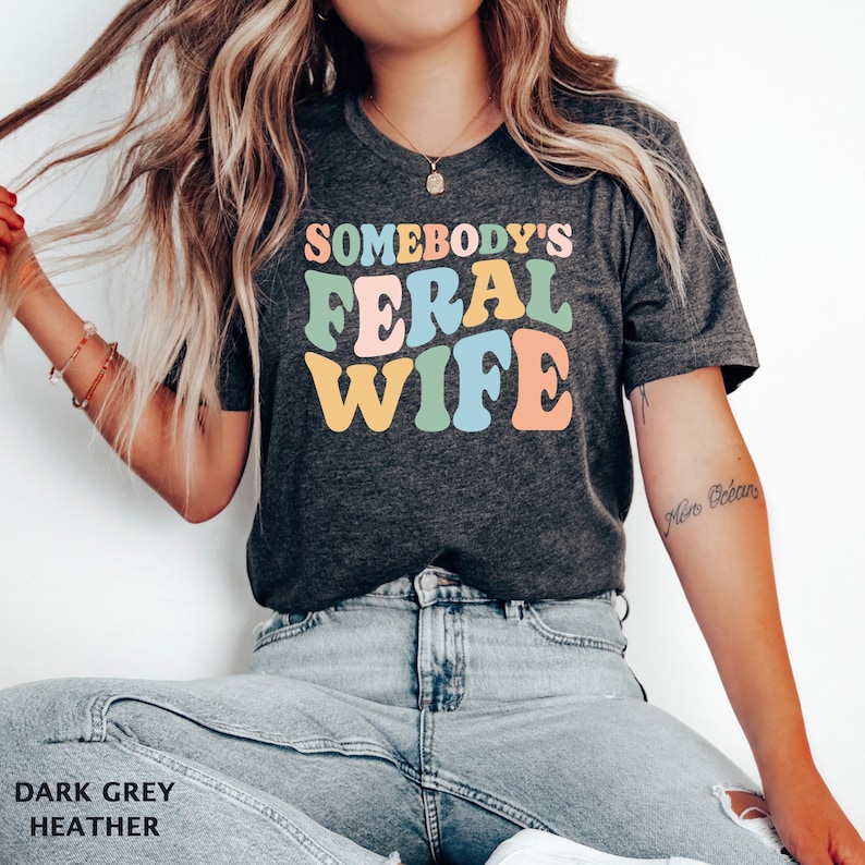 May include: A dark grey heather t-shirt with the text "Somebody's Feral Wife" in colorful retro lettering.