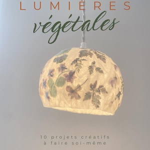 Op de afbeelding: Hanglamp met bloemenmotief. De crèmekleurige lampenkap is bolvormig en versierd met paarse, groene en witte bloemenpatronen. De tekst "LUMIÈRES végétales" staat bovenaan en "10 projets créatifs à faire soi-même" onderaan.