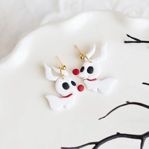 May include: A pair of white polymer clay earrings shaped like the character Zero from the movie The Nightmare Before Christmas. The earrings have black eyes, red noses, and a red collar.