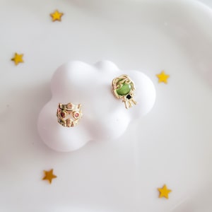 May include: Two gold-colored metal charms on a white cloud-shaped surface. One charm is a small crown with red accents. The other is a figure with a green face and black details. Scattered around are small gold star-shaped decorations.
