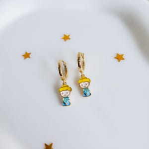 May include: A pair of gold-colored hoop earrings with a cartoon character charm. The charm features a yellow hat, white face, blue dress, and a gold-colored hoop. The earrings are on a white surface with gold star-shaped confetti.