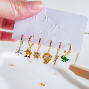 Può includere: Un set di sei orecchini a cerchio color oro con vari ciondoli. I ciondoli includono un fiore viola, un fiore giallo, un sole, un personaggio dei cartoni animati, un pesce e un dinosauro verde. Il testo "ROSA Diamante Accessories" è visibile.