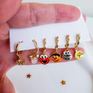 Pode incluir: Um conjunto de seis brincos de argola dourados com vários pingentes. Os pingentes incluem uma estrela, uma gema rosa e rostos de personagens de desenhos animados em vermelho, laranja, azul e amarelo. Os brincos são exibidos em um cartão branco.