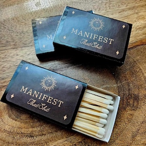 May include: Two black matchboxes with a gold sun and moon design and the text "MANIFEST That Shit". The matchboxes are open and filled with wooden matches.