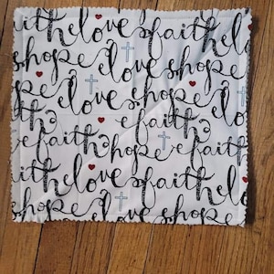May include: White fabric with black cursive writing that says "with the love of faith the love hope love faith the love of faith the love hope love" with small red hearts and crosses throughout.