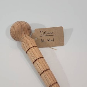 May include: A wooden dibber made of ash wood. The dibber has a rounded top and a cylindrical body with several carved rings. A tag attached to the dibber reads "Dibber Ash Wood".