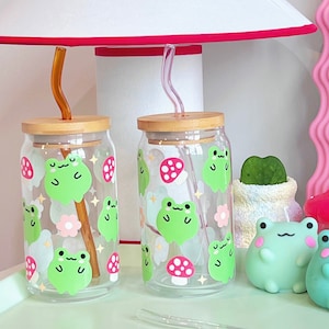 May include: Two clear glass reusable drinking cups with bamboo lids and straws. The cups have a frog and mushroom design with pastel colors.