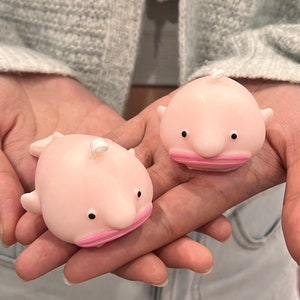 Op de afbeelding: Twee roze rubberen squish-speelgoed in de vorm van een anglerfish met roze monden en zwarte ogen.