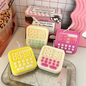 Puede incluir: Cuatro velas de temporizador de cocina con forma de relojes digitales. Las velas son amarillas, blancas y rosas. Cada vela tiene una pantalla digital que muestra "88:88" y tiene botones etiquetados como "Detener" e "Iniciar".