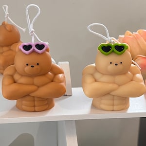 Peut inclure: Deux bougies en forme d'ours brun avec des muscles sculptés et portant des lunettes de soleil en forme de cœur. Une bougie a des lunettes de soleil roses et l'autre a des lunettes de soleil vertes.