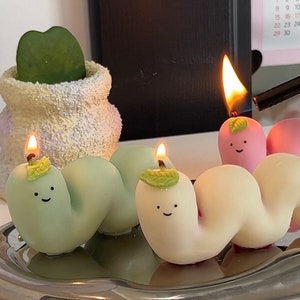 May include: Three lit candles shaped like green, white, and pink worms with happy faces. The candles are on a silver tray.