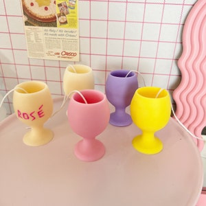 May include: Five votive candles in the shape of wine glasses. The candles are in various colors, including yellow, pink, purple, and cream. One candle has the word "ROSÉ" written on it.