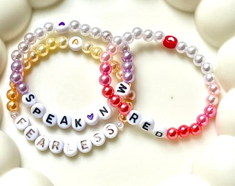 May include: Three beaded bracelets with white, gold, pink, and red beads. One bracelet spells "SPEAK" and "FEARLESS" with white letter beads. The other bracelets have the letters "A", "N", and "RED" with heart-shaped beads.