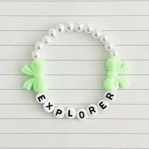 Könnte beinhalten: Ein weißes Perlenarmband mit grünen Schleifenanhängern und weißen Buchstabenperlen, die "EXPLORER" buchstabieren.