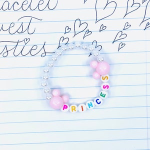 May include: A white beaded bracelet with pink and white beads spelling out the word "Princess" in colorful letters.