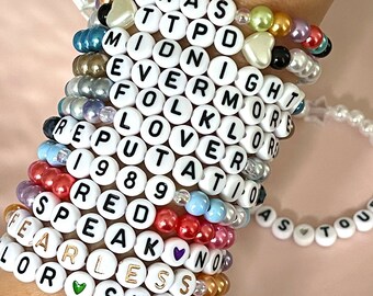 May include: A collection of beaded bracelets spelling out song titles and album names. The bracelets feature white beads with black letters, and colorful accent beads. The text includes "ERAS", "MIDNIGHT", "EVERMORE", "FOLKLORE", "LOVER", "REPUTATION", "1989", "RED", "SPEAK NOW", "FEARLESS", and "SWIFT".