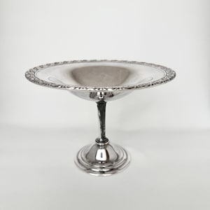 May include: A silver-colored, pedestal serving bowl with a decorative, scalloped rim. The bowl is supported by a slender, textured stem and a circular base. The bowl is likely used for serving fruit or desserts.