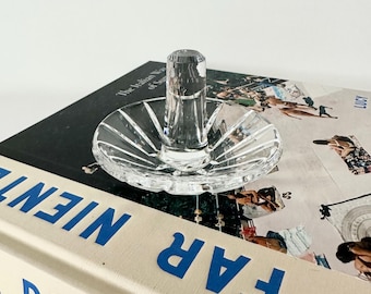 May include: A clear, cut-glass ring holder with a circular base and a central post. The base has a starburst design. The ring holder is resting on a book with the words "FAR NIENTE DOLCE" in blue letters.