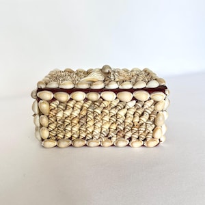 May include: A rectangular box covered in various small, cream-colored seashells. The lid and sides are densely decorated with rows of spiraled and smooth shells, creating a textured surface over the dark brown base of the container.