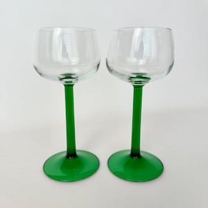 May include: Two clear wine glasses with green stems and bases. The glasses have a rounded bowl shape and a long, slender stem. The green color of the stem and base provides a striking contrast to the clear glass bowl.