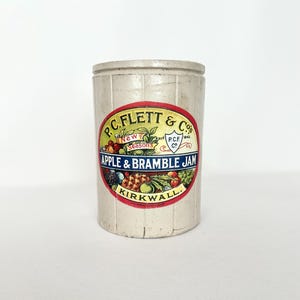 May include: A vintage, cylindrical stoneware jar with a cream-colored exterior. The jar features a colorful label with the text "P.C. Flett & Co" and "Apple & Bramble Jam, Kirkwall." The label includes illustrations of fruits and foliage.