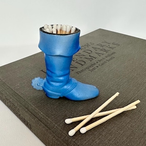 May include: Blue ceramic boot match holder with white tipped matches inside. Three additional matches are on a dark gray book with the text "World's Legendary Landmarks" on the cover.