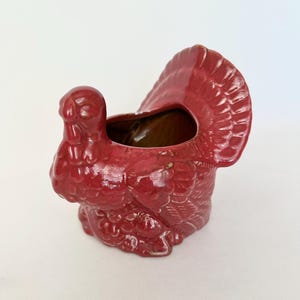 May include: A ceramic turkey-shaped planter in a glossy red finish. The planter has a detailed design, including feathers and a small opening at the top. The interior is a contrasting brown color. A festive Thanksgiving decoration.