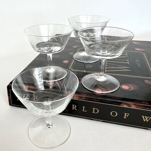 May include: Four clear champagne coupe glasses with etched leaf designs. The glasses have a wide, shallow bowl and a long stem. They are arranged on a dark book with gold lettering. The glasses are made of glass.