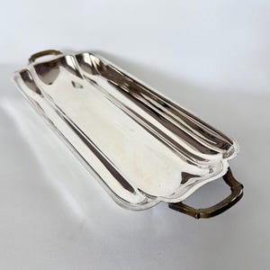 May include: A rectangular silver-toned serving tray with a polished, reflective surface and scalloped edges. The tray features two dark, metallic handles on opposite ends, each with a curved, ornate design that contrasts with the smooth body.