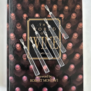 May include: A hardcover book titled "The World of Wine" with a background of wine bottles. Six clear, decorative forks with silver tines are arranged on top. The book's cover features gold lettering and the foreword is by Robert Mondavi.