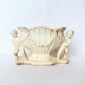 May include: White ceramic planter shaped like a seashell with two cherub figures on either side.