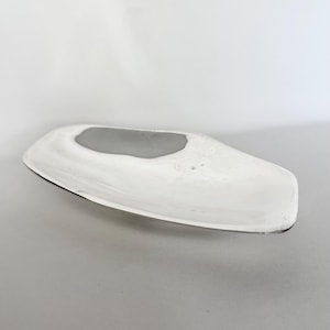 May include: A polished silver-toned metal tray with an elongated, irregular, and organic shape. The surface is smooth and reflective, featuring a shallow, curved basin that tapers toward the ends, creating a sleek, modern, and minimalist aesthetic.