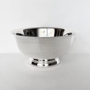 May include: A polished silver-toned bowl with a smooth, reflective surface and a wide, open top. The bowl features a circular, tiered pedestal base that provides stability, with a clean, minimalist design and a consistent metallic finish throughout.