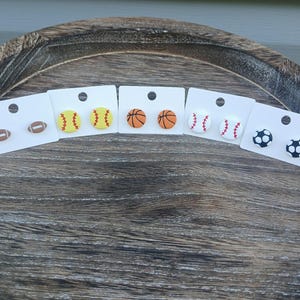 May include: A collection of sports-themed stud earrings. The earrings feature designs of a football, baseball, basketball, soccer ball, and softball. Each pair is mounted on a white card with a black stud post.