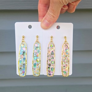 May include: Four pairs of dangle earrings with gold-colored frames. Two pairs have a light blue background, and two have a pink background. Each earring is filled with iridescent abalone shell pieces.