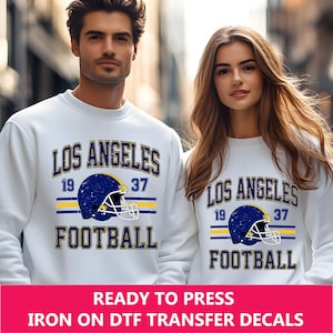 May include: White sweatshirt with a blue and yellow graphic design that says "Los Angeles Football" with a football helmet graphic. The graphic has a distressed look.