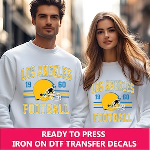 May include: White sweatshirt with a blue and yellow Los Angeles Football design. The design includes the text "Los Angeles" and "Football" with the numbers "19" and "60" on either side of a football helmet.