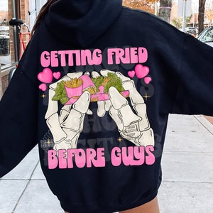 Op de afbeelding: Zwarte hoodie met de roze tekst "GETTING FRIED BEFORE GUYS", met een afbeelding van skeletachtige handen die een bord friet en een taco vasthouden. Het ontwerp bevat roze harten en een cartoonachtige stijl.