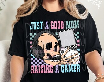 May include: Black t-shirt with a pink, blue, and white graphic of a skull wearing headphones and holding a video game controller. The text "Just a good mom raising a gamer" is printed on the shirt.