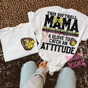 May include: White t-shirt with the text "This Softball Mama Don't Need A Glove To Catch An Attitude" in black with a yellow and red stripe and a yellow bow. The shirt also features a baseball glove with a yellow baseball and a yellow bow.