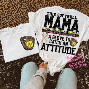 K&ouml;nnte beinhalten: Wei&szlig;es T-Shirt mit dem Text "This Softball Mama Don't Need A Glove To Catch An Attitude" in Schwarz und Rot. Das Shirt hat eine Grafik eines Baseballhandschuhs und eines Balls.