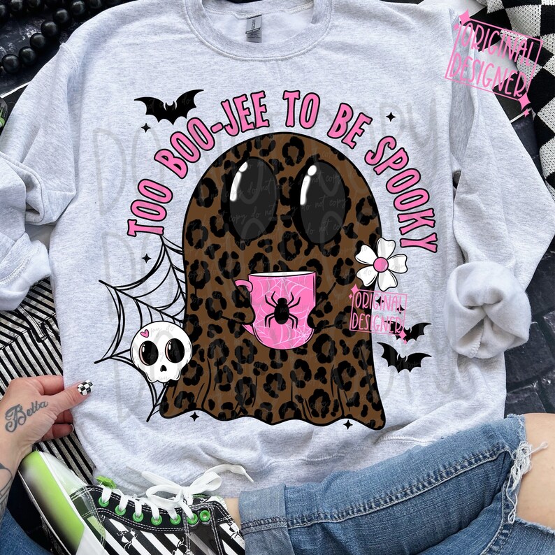 Op de afbeelding: Een lichtgrijze sweatshirt met een Halloween-thema. Het ontwerp toont een spook met een luipaardprint, die een roze mok met een spin vasthoudt. De tekst "TOO BOO-JEE TO BE SPOOKY" is roze. Vleermuizen, een spinnenweb en een schedel maken het ontwerp compleet.