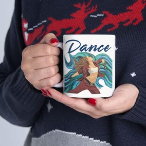 May include: White ceramic mug featuring the word "Dance" in a script font. The mug displays a dancer in a red outfit with blue and green flowing lines. The mug is held by a person wearing a blue sweater.