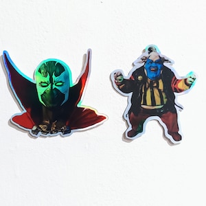 May include: Two holographic stickers featuring comic book characters. The first sticker is of a green and black masked character with a red cape. The second sticker is of a blue and yellow character with a red and yellow jacket.