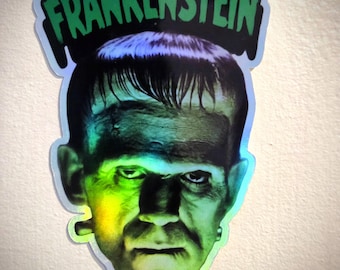 May include: A holographic sticker featuring a portrait of Frankenstein's monster. The monster's face is green and black, with the word "FRANKENSTEIN" in green above his head. The sticker has a glossy, iridescent finish.