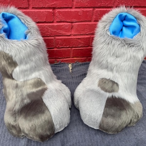 May include: A pair of furry gray and brown costume feet with blue interior lining. The faux fur feet have a two-tone design, with a lighter gray on top and a darker gray on the bottom. The feet are designed to be worn as part of a costume.