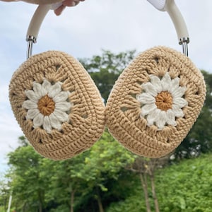 Puede incluir: Calentadores de orejas de crochet con un patrón de margarita en beige y blanco. Los calentadores de orejas están unidos a una diadema.