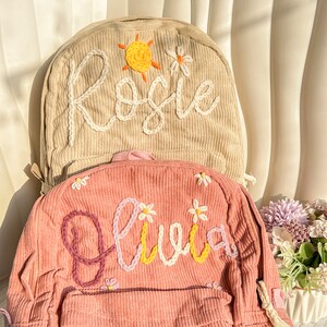 May include: Two corduroy backpacks, one beige and one pink, are shown. The beige backpack has the name "Rosie" embroidered in white, with a sun and flower design. The pink backpack features the name "Olivia" in various colours and flower details.