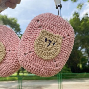 Pu&ograve; includere: Copri cuffie in crochet rosa con un viso di orso in crochet marrone su un lato. Il viso dell'orso ha un'espressione di accigliamento ricamata in nero.