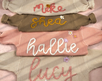 May include: Four knitted sweaters stacked on top of each other. The sweaters are beige, brown, pink, and beige. Each sweater has a name embroidered on it: "Mike", "Shea", "Hallie", and "Lucy".